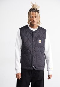 Black quilted vest with a stand-up collar, snap buttons, and a front pocket, worn over a gray sweatshirt, paired with dark pants.