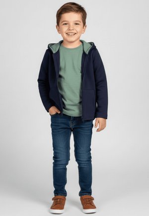 Uśmiechnięty młody chłopiec o brązowych włosach, ubrany w granatową bluzę z kapturem, zieloną koszulkę, niebieskie dżinsy i brązowe buty, stoi z jedną ręką w kieszeni.