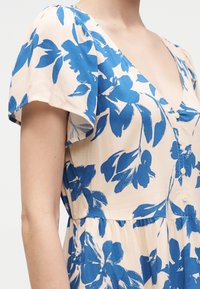 Φόρεμα κρεμ με μπλε floral σχέδιο, κοντά φτερωτά μανίκια, V-λαιμόκοψη και κουμπιά μπροστά, φορεμένο στο πάνω μέρος του σώματος και το χέρι.