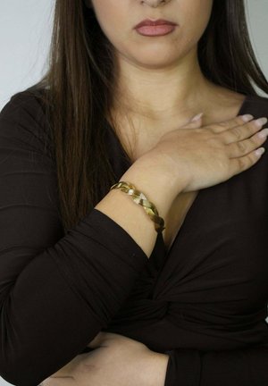 Złota, skręcana metalowa bransoletka noszona na nadgarstku, z teksturowanym wzorem. Osoba nosząca bransoletkę ma długie, brązowe włosy i ciemną bluzkę.