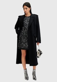Cappotto nero sopra un vestito nero decorato con paillettes, abbinato a stivali metallici alla caviglia e una borsa argentata. Tessuto morbido e design su misura.