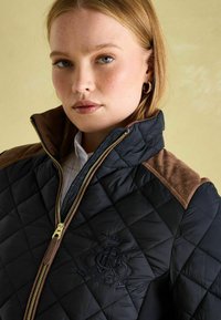 Jachetă neagră matlasată, cu accente din piele întoarsă maro pe umeri, fermoar auriu și logo brodat. Textură netedă, cu elemente de design funcționale.