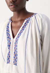 Gros plan d'une personne portant un chemisier écru avec une broderie géométrique bleue et rouge le long de l'encolure et des manches.