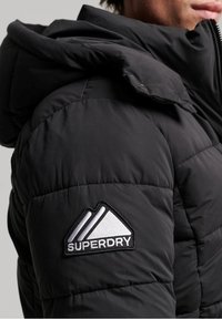 Czarna kurtka puchowa z kapturem, z pikowanymi sekcjami i wyraźnym haftowanym logo Superdry na rękawie. Gładka tekstura.