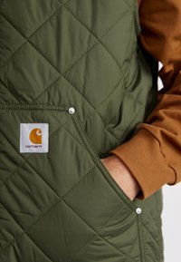 Zeleno prešito brezrokavnik z žepom na zapenjanje in oznako Carhartt. Svetlo rjava manšeta na rokavu je vidna in prikazuje kontrastno barvo.