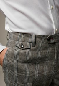 Grau karierte Hose mit blauen und schwarzen Streifen. Der Stoff hat eine glatte Struktur und verfügt über eine seitliche Tasche mit einem schwarzen Knopf, kombiniert mit einem weißen Hemd.
