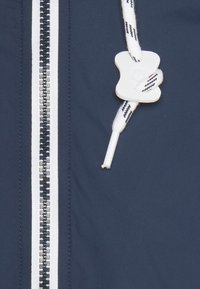 Giacca blu navy con zip argentata, cordino bianco con accento nero e un bottone di chiusura bianco. Texture liscia e design lineare.