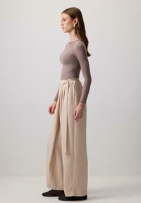 Woman standing in profile wearing a fitted mauve long-sleeve top, high-waisted beige wide-leg pants with belt, and black shoes.