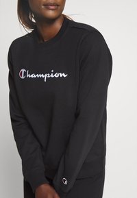 Sweatshirt noir en coton avec un logo "Champion" brodé en blanc et rouge. Poignets et ourlet côtelés. Design simple.
