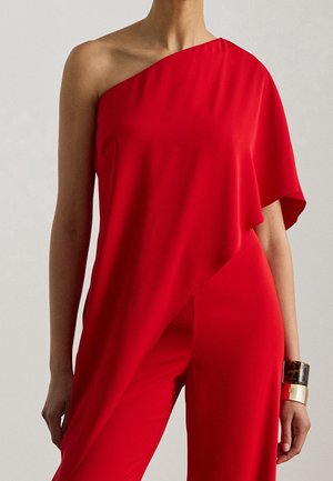 Femme portant un top rouge vif asymétrique et un pantalon rouge assorti avec deux larges bracelets à son poignet droit.