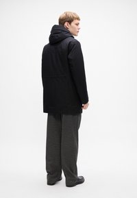 Jonge man die staat, gekleed in een zwarte capuchonjas, grijze wijde broek en zwarte schoenen, die zijwaarts kijkt tegen een witte achtergrond.