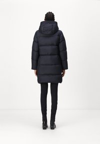 Tummansininen untuvatakki hupulla, tikattu malli ja resorit hihansuissa, yhdistettynä kapeisiin mustiin housuihin ja korollisiin nilkkureihin.