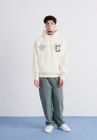 Sudadera crema con bolsillo frontal, que presenta texto gráfico en negro y un logo. Combinada con pantalones sueltos de color verde oliva y zapatillas blancas.