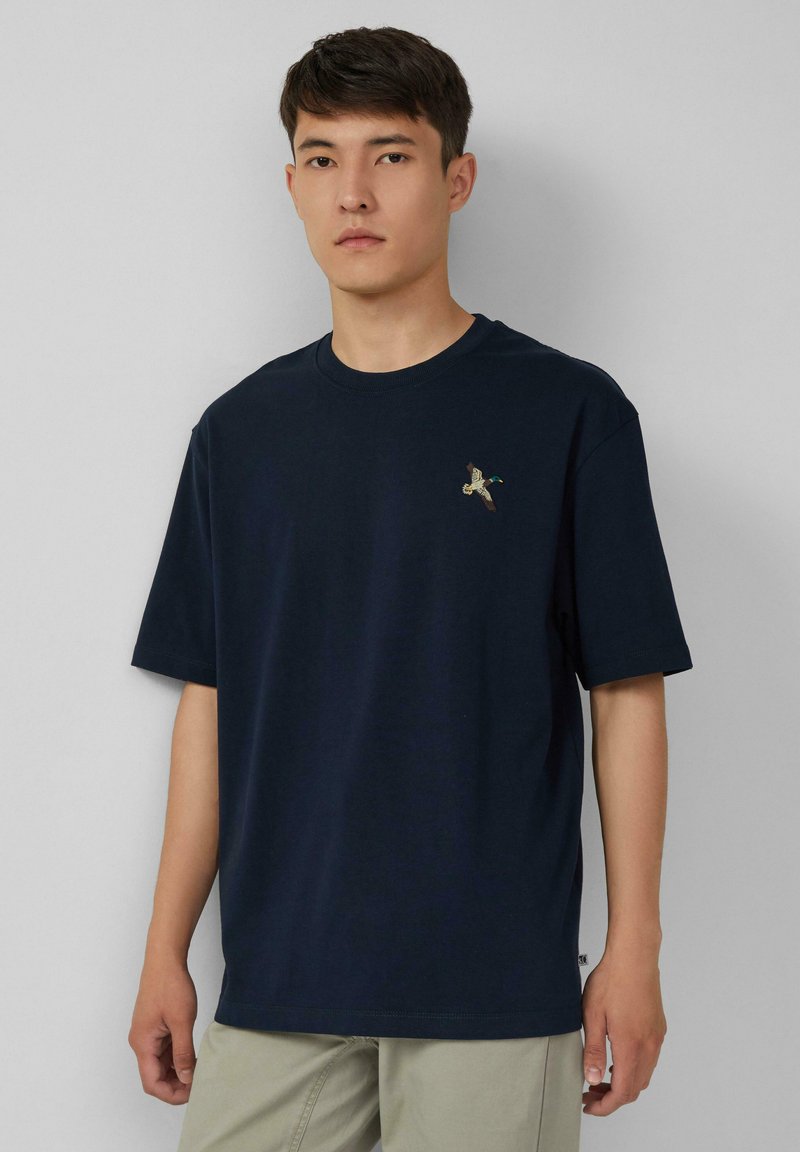 Junger Mann in einem navyblauen T-Shirt mit einem kleinen gestickten fliegenden Entenmotiv auf der linken Brust und beigen Hosen, der vor einer hellgrauen Wand steht.
