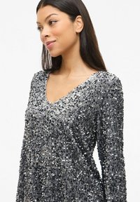 Sudraba krāsas sequined tops ar V-veida izgriezumu un garām piedurknēm. Rakstā ir teksturēta virsma ar metālisku spīdumu, apvienojot pelēktoņu toņus dizainā.