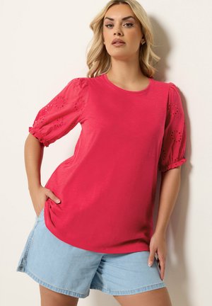 Kvinde iført en kraftigt pink bluse med puffede, hulmønstrede ærmer og lyseblå denimshorts, poserer mod en ensfarvet baggrund.