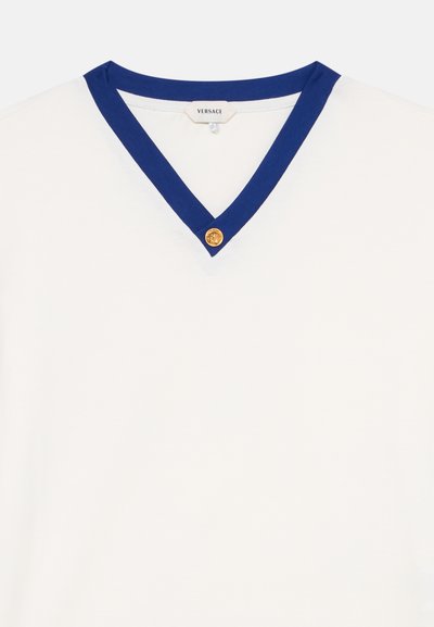 T-shirt blanc à col en V avec un liseré bleu comportant un bouton médaillon doré au centre et une étiquette Versace visible à l’intérieur du col.