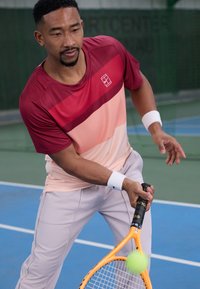 Rød og ferskenfarget gradient treningsskjorte, lys grå bukser, hvite håndleddbånd, holder et oransje tennisracket med en grønn tennisball i bevegelse.