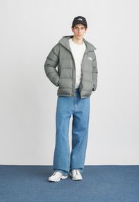 Giacca imbottita in grigio tenue, con cappuccio e logo sul petto; abbinata a jeans larghi blu e sneakers bianche. Un cappellino nero completa il look.