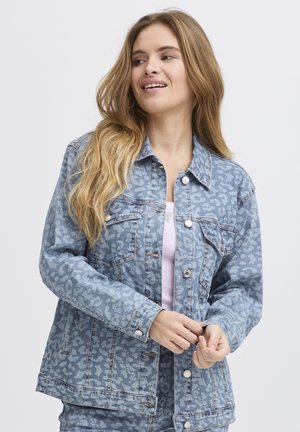 Giovane donna con lunghi capelli biondi che indossa una giacca di jeans azzurro chiaro con motivo sopra una maglia bianca, guarda a sinistra e sorride leggermente.