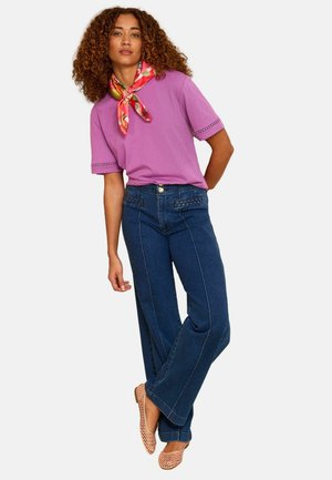 Nainen, jolla on kiharat hiukset ja päällään violetti lyhythihainen paita, tummansiniset farkut, kukallinen kaulahuivi ja beige punottu kengät, seisoo jalat ristissä.