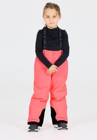 Leuchtend pinke Schneehosen mit schwarzen Akzenten, verstellbaren Trägern und einer lockeren Passform, kombiniert mit einem schwarzen Langarmoberteil und sportlichen Schuhen.