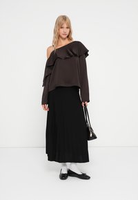 Top marrone con spalle scoperte e dettagli arricciati, abbinato a una gonna maxi nera plissettata. La modella tiene in mano una borsa nera, indossa calzini bianchi e ballerine nere.