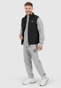 Černá prošívaná vesta s vysokým límcem, pod ní šedá mikina a odpovídající tepláky. Na veste je logo a na kalhotách emblém. Bílé tenisky.