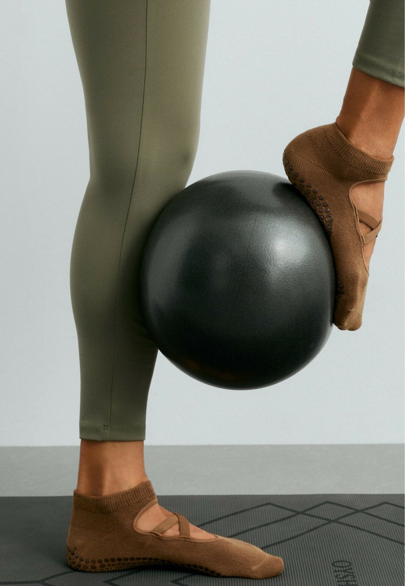 Jambes en leggings olive et chaussettes antidérapantes marron équilibrant un ballon d'exercice noir entre les pieds sur un tapis d'exercice noir.