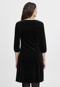 Schwarzes Samtkleid mit tailliertem Oberteil, dreiviertellangen Ärmeln und einem ausgestellten Rock. Es hat eine glatte Textur und einen subtilen Glanz.