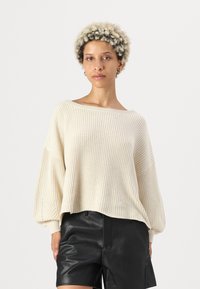 Cremefarbener, gestrickter Pullover mit weitem Ausschnitt und lässiger Passform, der über eine gerippte Textur und Ballonärmel verfügt, kombiniert mit schwarzen Leder-Shorts.