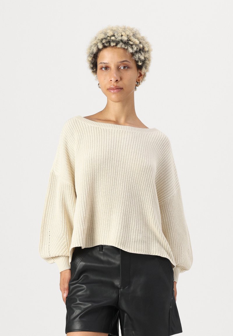 Cremefarbener, gestrickter Pullover mit weitem Ausschnitt und lässiger Passform, der über eine gerippte Textur und Ballonärmel verfügt, kombiniert mit schwarzen Leder-Shorts.