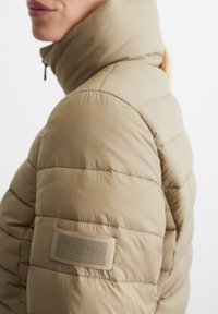 Beige gevoerde jas met een hoge kraag, stiksel details en een merkpatch op de linkermouw. Ritssluiting. Glad textuur.