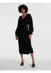 Schwarzes Wickelkleid mit langen Puffärmeln, mit Knopfdetails an der Seite und einem knielangen Schnitt, aus einem leichten Stoff gefertigt.