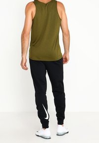 Débardeur sans manches vert olive associé à un pantalon noir arborant un logo Nike blanc et des baskets blanches à semelle texturée.