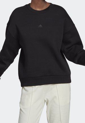 Personne portant un sweat-shirt Adidas noir avec un petit logo sur la poitrine et un pantalon blanc ample sur un fond uni.