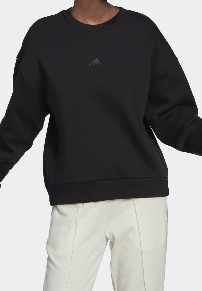 Personne portant un sweat-shirt Adidas noir avec un petit logo sur la poitrine et un pantalon blanc ample sur un fond uni.