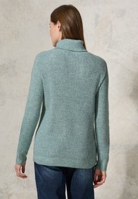 Hellgrüner Strickpullover mit Rollkragen, strukturiertem Muster, geripptem Saum und langen Ärmeln, kombiniert mit dunkelblauen Jeans.