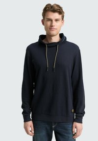 Navy blauwe pullover hoodie met een kraag, gouden trekkoorden, geribbelde textuur en een kleine accentpatch aan de onderkant. Lange mouwen.