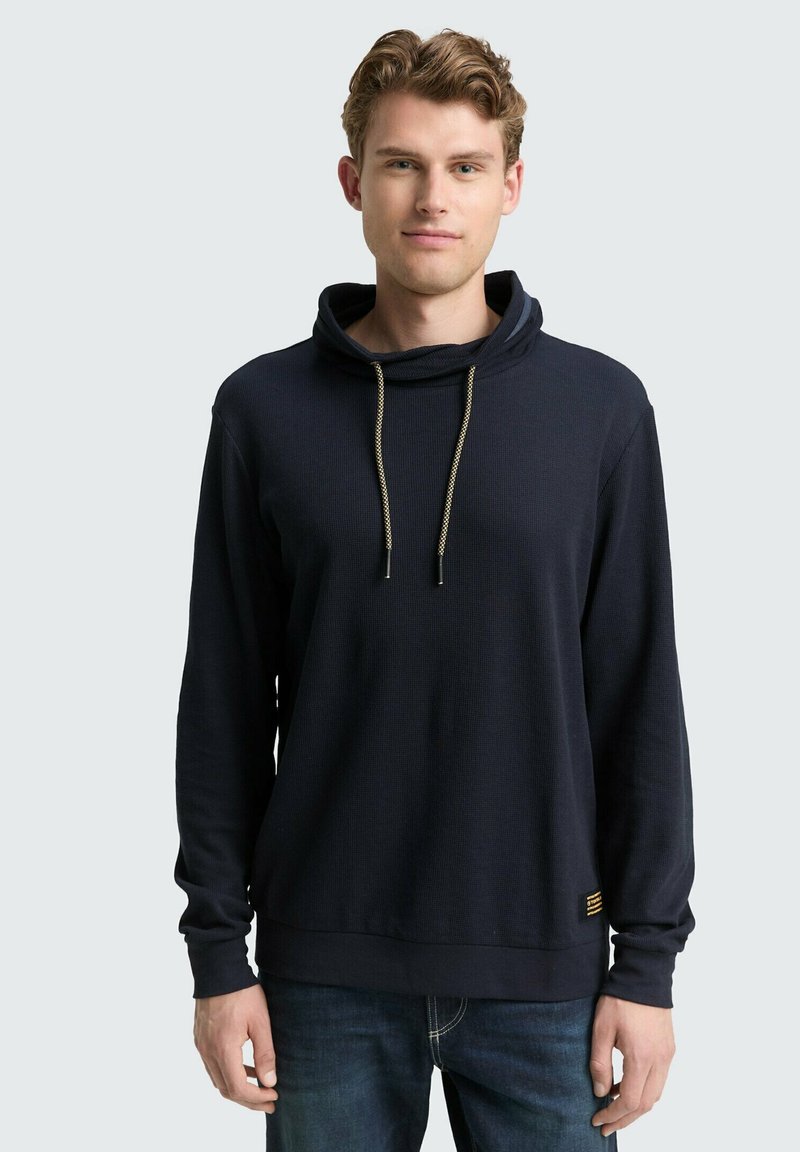 Navy blauwe pullover hoodie met een kraag, gouden trekkoorden, geribbelde textuur en een kleine accentpatch aan de onderkant. Lange mouwen.