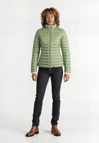 Lichtgroene, gewatteerde jas met een hoge kraag, ritssluiting aan de voorkant en een oranje accent. Draagt met zwarte broek en bruine enkellaarzen.