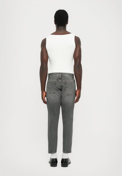 Man standing facing away, wearing a white sleeveless top, gray jeans, white socks, and black shoes against a plain white background.