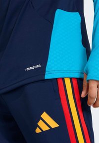 Top atlético azul-marinho com detalhes em turquesa, apresentando um design texturizado. Logo amarelo da Adidas nas calças com riscas laterais vermelhas e amarelas.