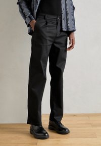 Čierne nohavice vyrobené z hladkej tkaniny s rovnými nohavicami, s predným gombíkom a vreckami, doplnené o elegantné čierne topánky.