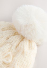 Bonnet tricoté de couleur crème avec un motif texturé, doté d'un grand pompon blanc et duveteux sur le dessus. Matière douce et tactile.