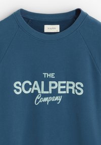 Tegen een teal sweatshirt met raglanmouwen, met op de voorkant witte letters "THE SCALPERS Company", gemaakt van zacht materiaal met een geribbelde kraag.