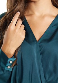 Femme portant un chemisier en satin teal avec des boutons en cristal sur les poignets, touchant ses longs cheveux bruns et portant une bague en or à son doigt.