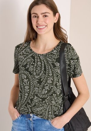 Jeune femme portant un haut à manches courtes à motif paisley vert, un jean bleu, et portant un sac bandoulière noir matelassé.
