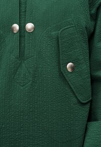 Υφασμα σκούρου πράσινου με υφή, με δύο μεγάλα στρογγυλά ασημένια κουμπιά κοντά στον γιακά και μια τσέπη με καπάκι που ασφαλίζει με ένα ασημένιο κουμπί ταιριαστό.