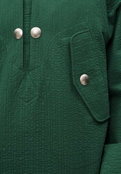 Υφασμα σκούρου πράσινου με υφή, με δύο μεγάλα στρογγυλά ασημένια κουμπιά κοντά στον γιακά και μια τσέπη με καπάκι που ασφαλίζει με ένα ασημένιο κουμπί ταιριαστό.